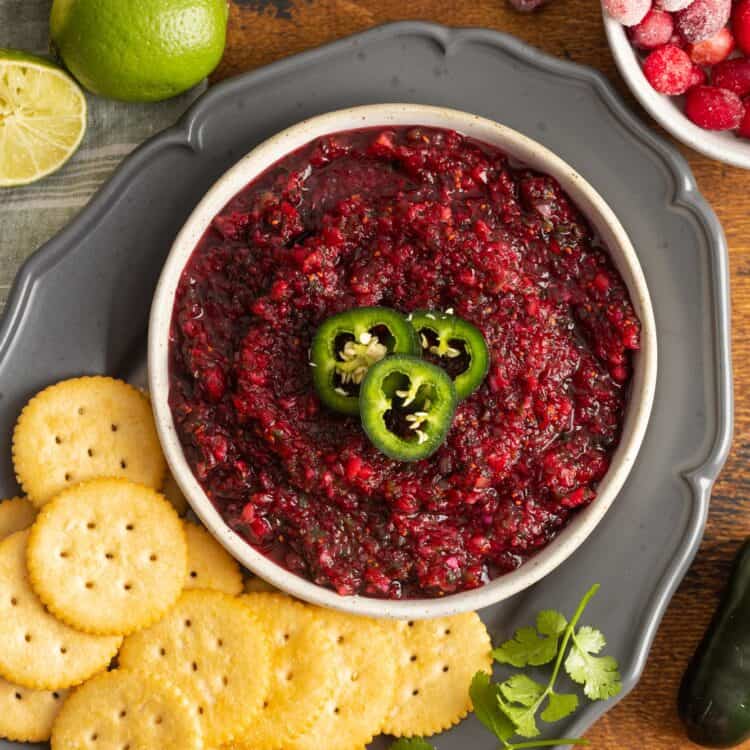 overhead view of a bowl of cranberry salsa, topped with sliced jalapenos, on a tray with ritz crackers and cilantro.