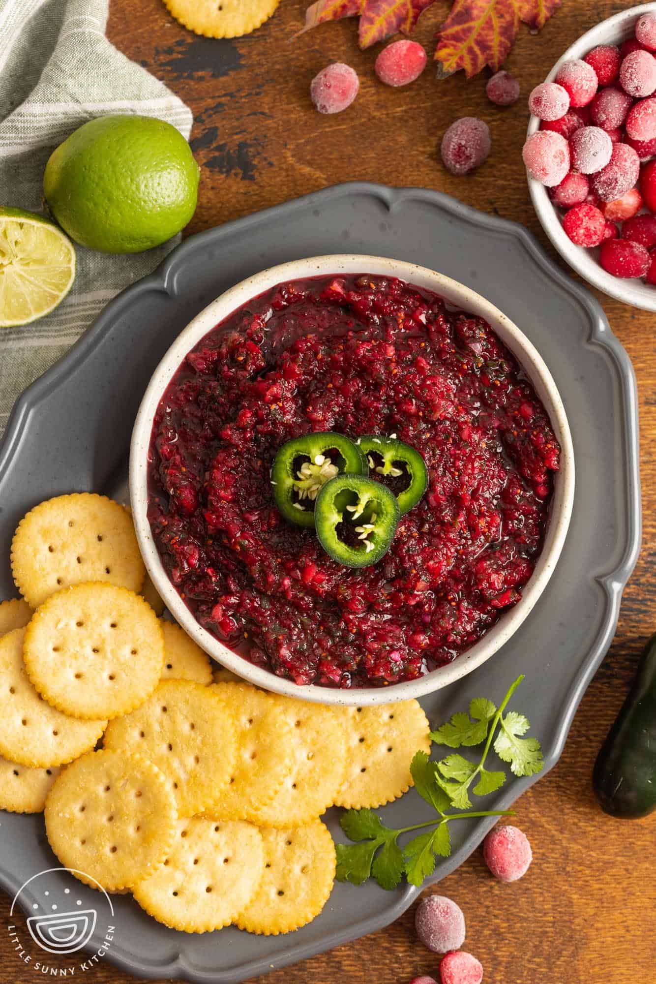 overhead view of a bowl of cranberry salsa, topped with sliced jalapenos, on a tray with ritz crackers and cilantro.