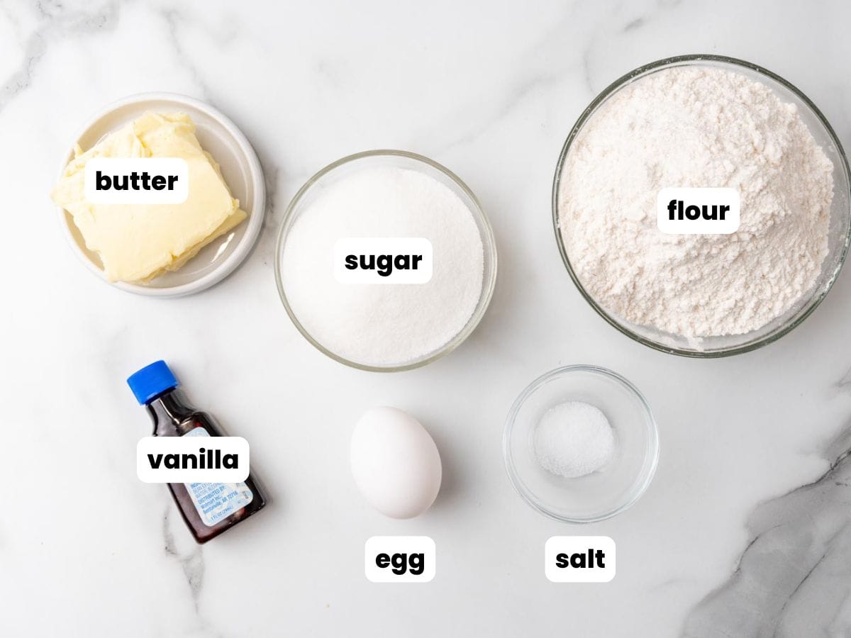 The six ingredients needed to make danish butter cookies: butter, sugar, flour, vanilla, egg, salt, all in separate bowls on a marble counters.