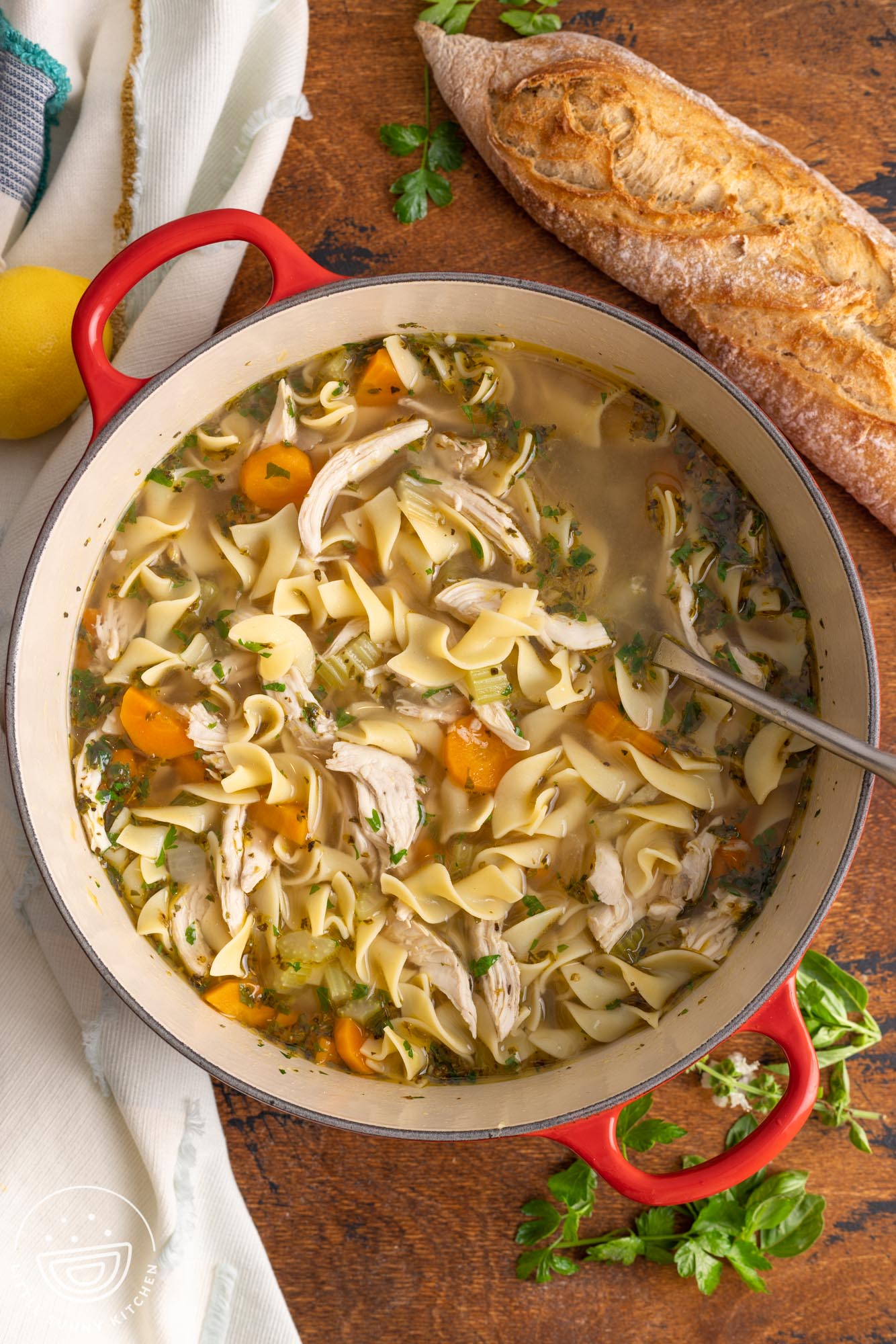 a red handled dutch oven of chicken soup with egg noodles, garnished with parsley, sitting on a wooden board.