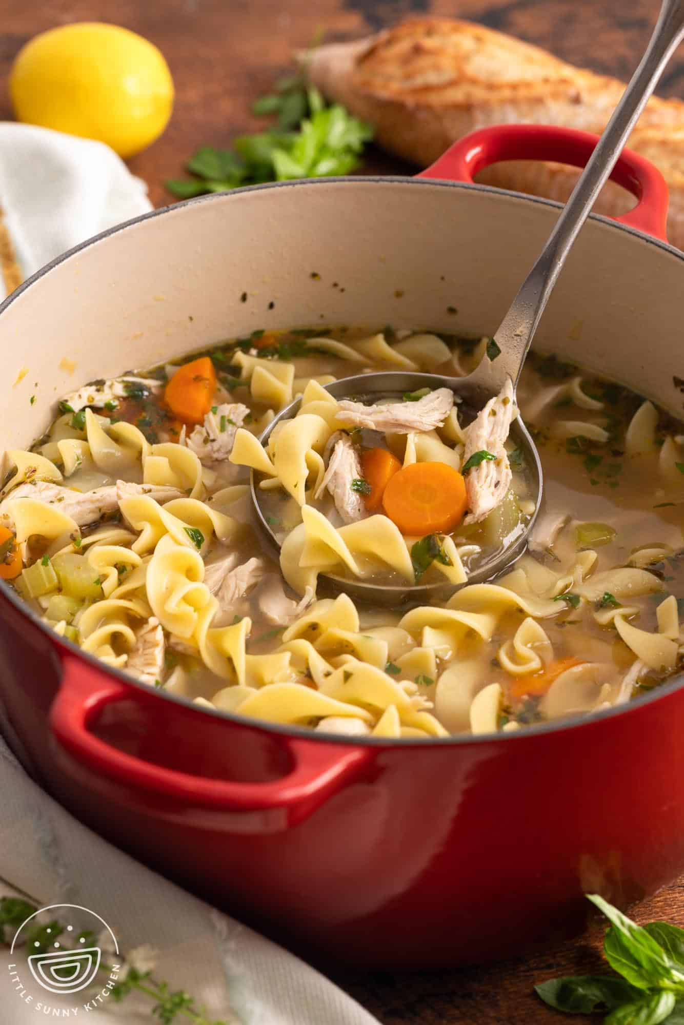a red dutch oven filled with hearty chicken noodle soup, served with a ladle. In the background is fresh herbs, a lemon, and a loaf of bread.