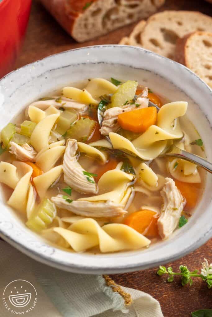 A bowl of chicken noodle soup with egg noodles on a wooden board in front of a loaf of bread.