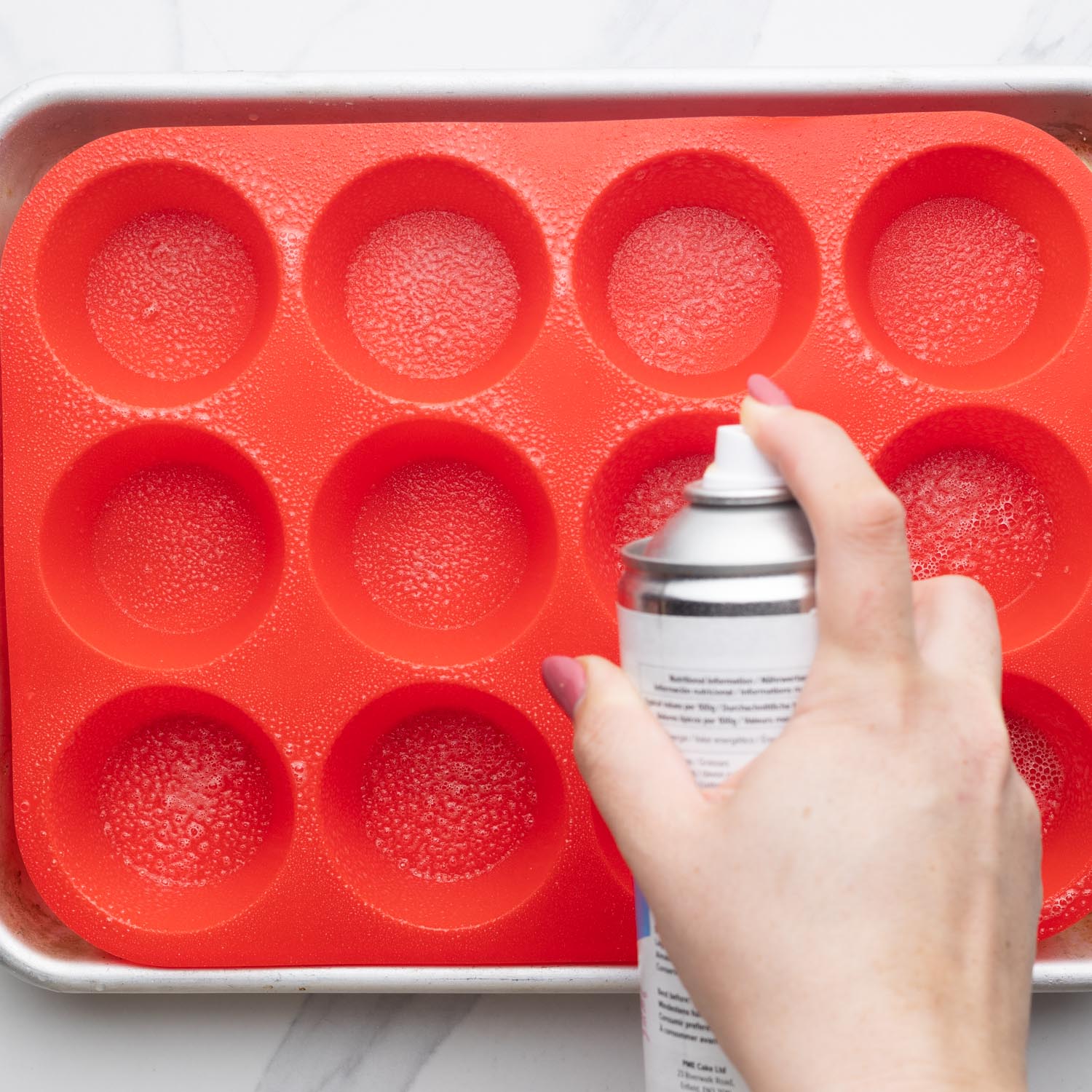 a hand using cooking spray to liberally grease a silicone muffin pan.