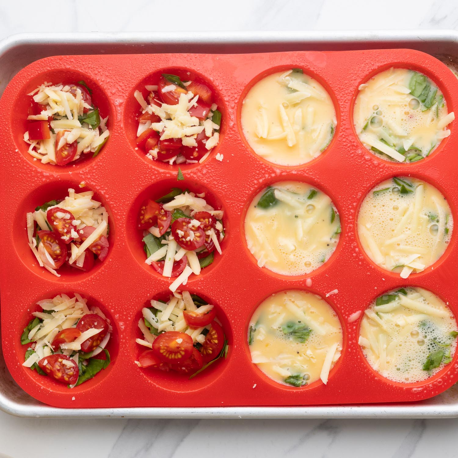 a red silicone muffin pan. 6 cavities are filled with shredded cheese, tomatoes, and spinach. The other six have been topped up with liquid eggs.