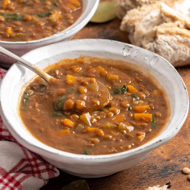 two white ceramic bowls of instant pot lentil soup with spinach and carrots. Around the bowls are pieces of crusty bread and bay leaves.