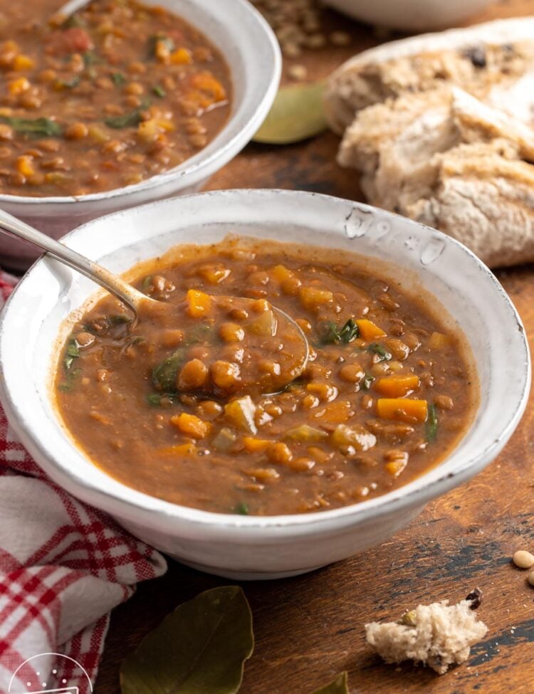 two white ceramic bowls of instant pot lentil soup with spinach and carrots. Around the bowls are pieces of crusty bread and bay leaves.