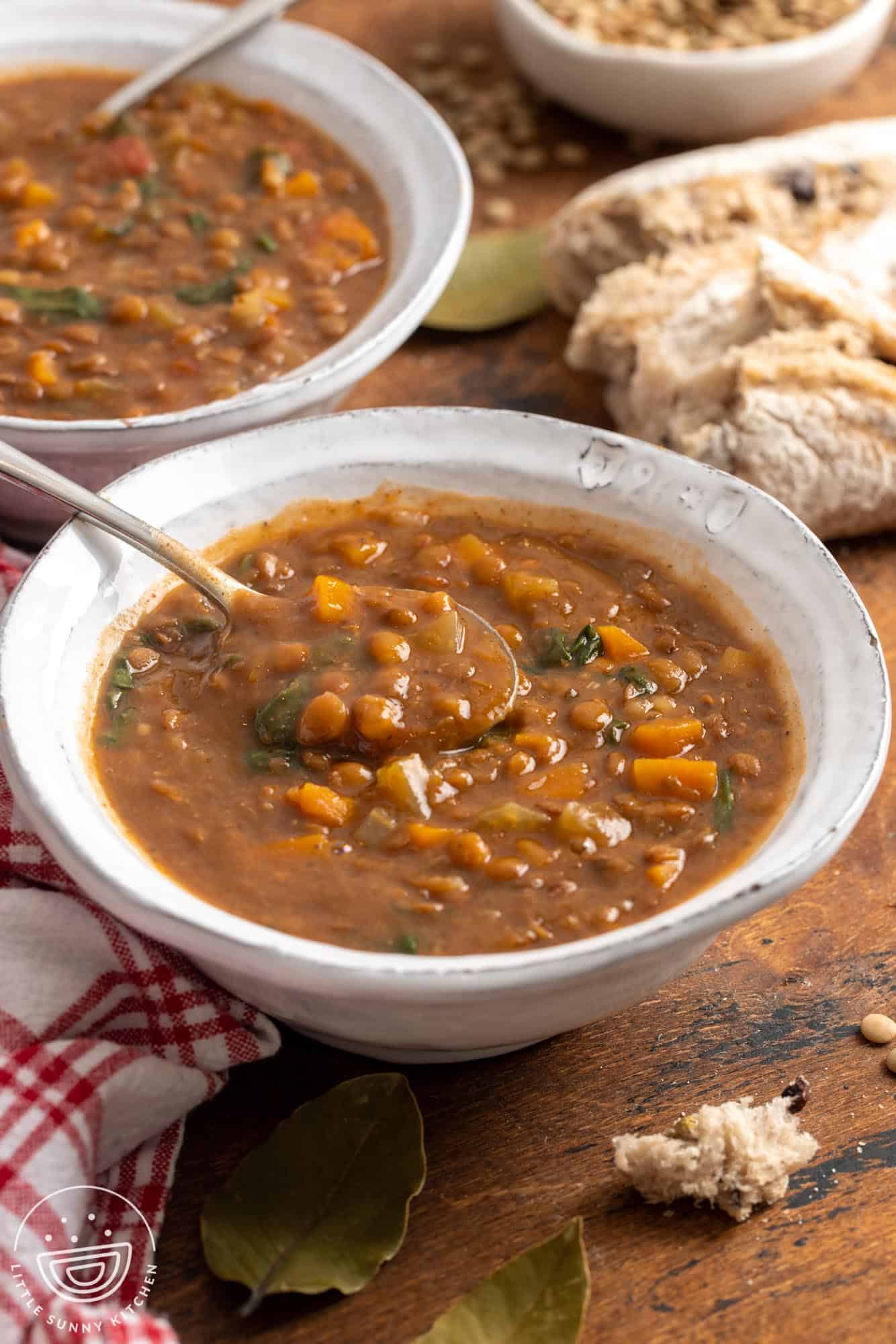 two white ceramic bowls of instant pot lentil soup with spinach and carrots. Around the bowls are pieces of crusty bread and bay leaves.