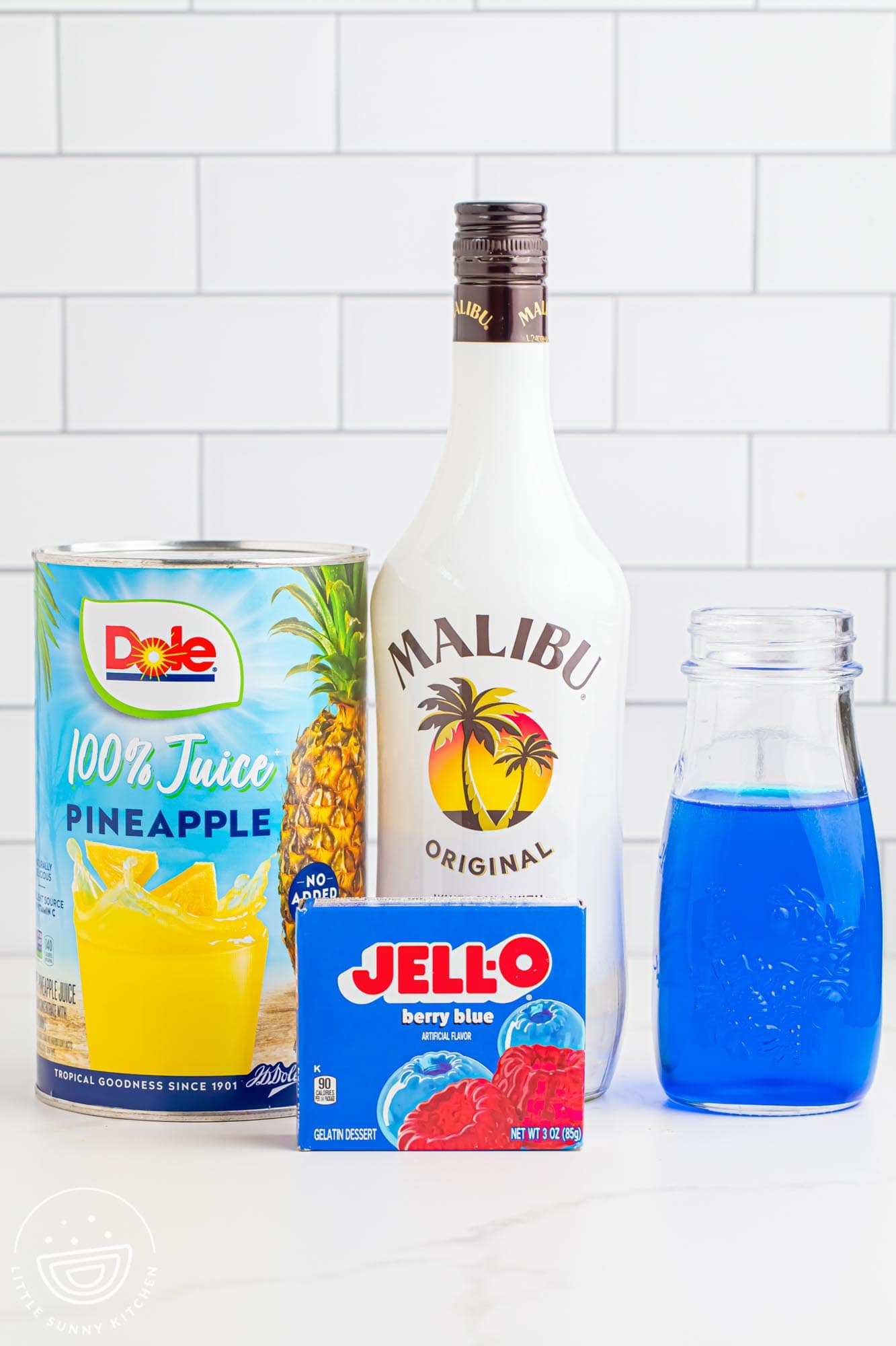 a bottle of malibu rum, a box of berry blue jello, a large can of pineapple juice, and a small glass bottle of blue curacao.