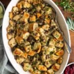 overhead view of an oval mushroom bread pudding casserole on a wooden table. Next to it is a bowl of cranberry sauce and a bowl of mashed squash.