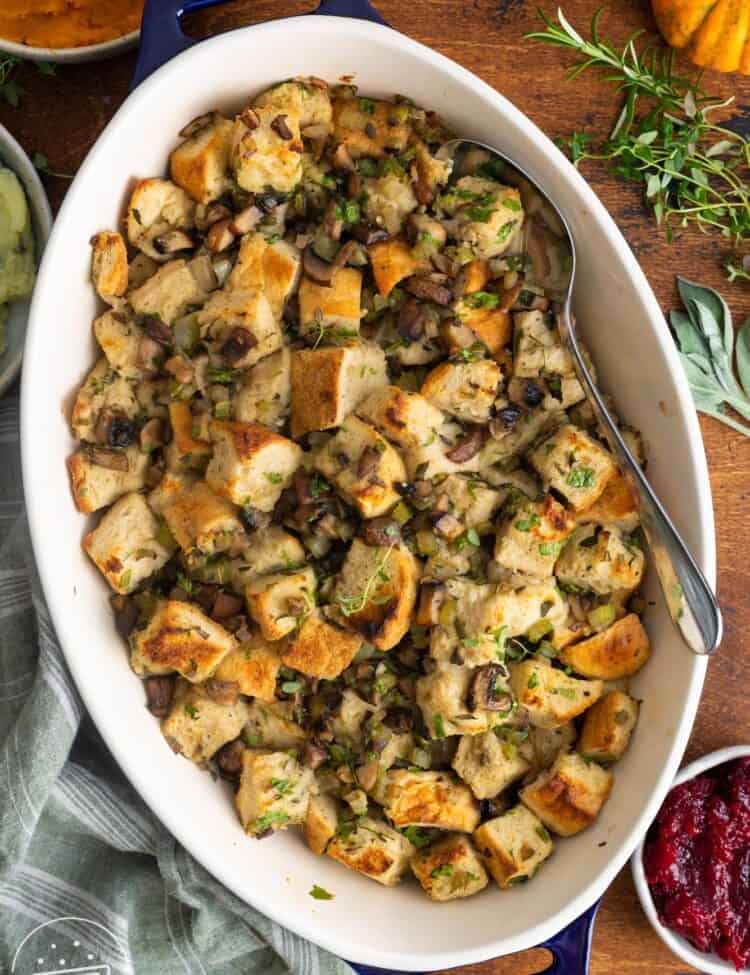 overhead view of an oval mushroom bread pudding casserole on a wooden table. Next to it is a bowl of cranberry sauce and a bowl of mashed squash.