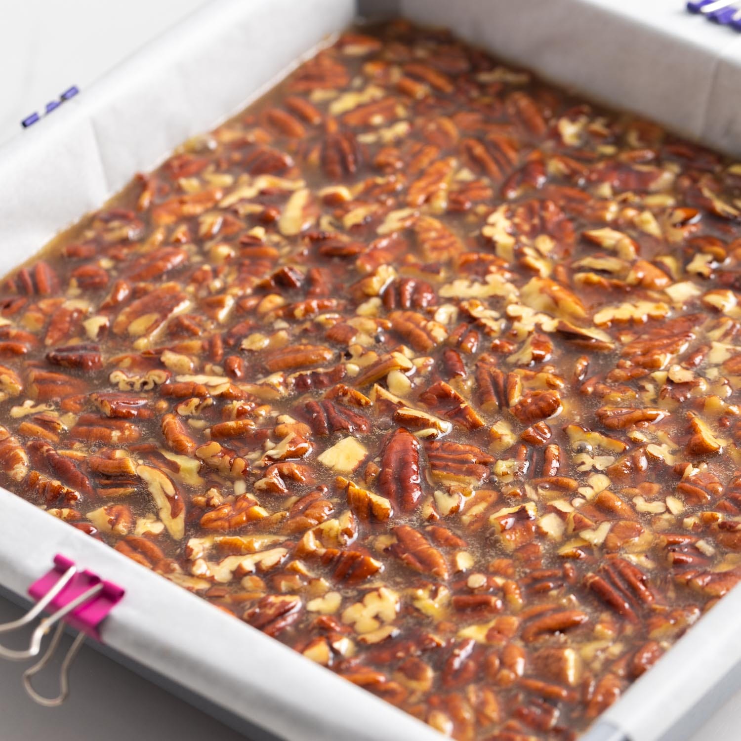 pecan pie bars in a square pan lined with parchment paper, before baking. The parchment paper is held down with purple binder clips.