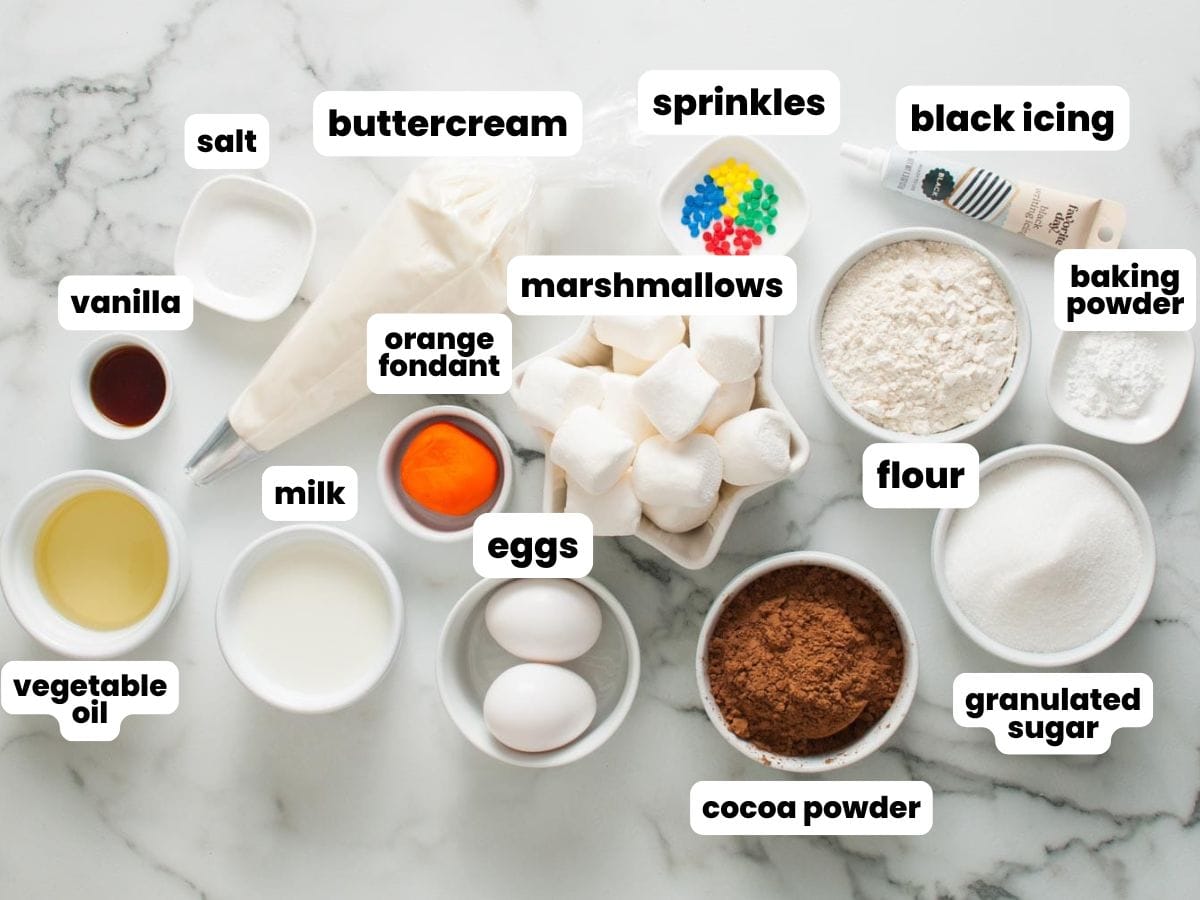 The ingredients needed to make chocolate cupcakes with marshmallow snowman decorations. All are measured in small white bowls and arranged on a marble counter.