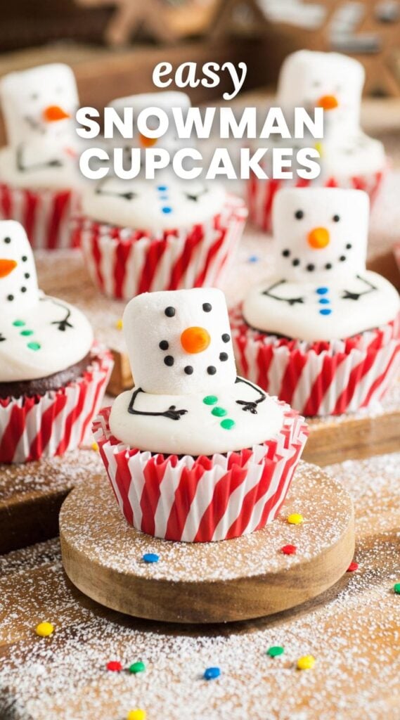 Chocolate cupcakes topped with melted marshmallow snowmen that are decorated with icing. The cupcakes are in red and white striped paper liners and resting on a wooden cutting board. Text overlay says "easy snowman cupcakes"