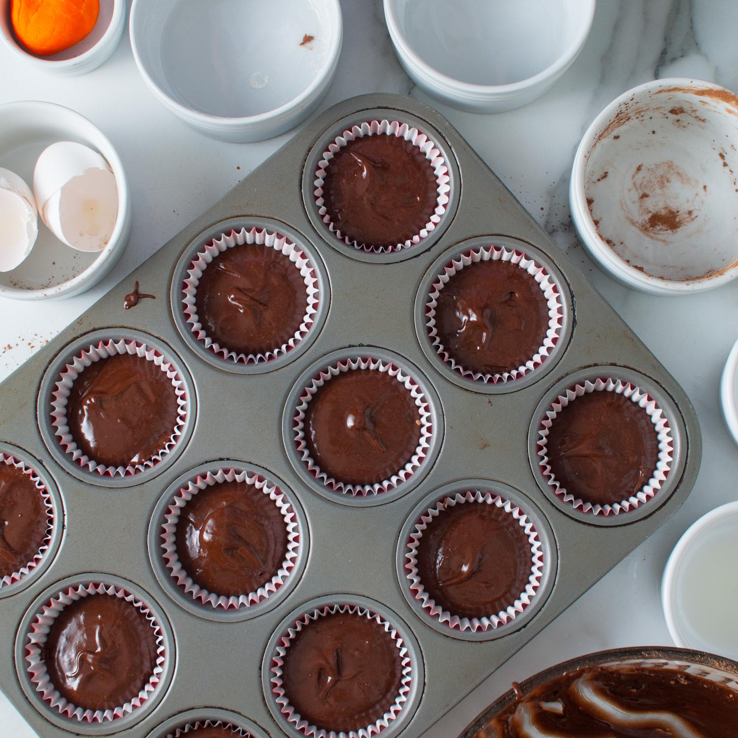 chocolate cupcake batter divided into 12 cupcake liners in a metal muffin tin. Around the tin are empty ingredient bowls.