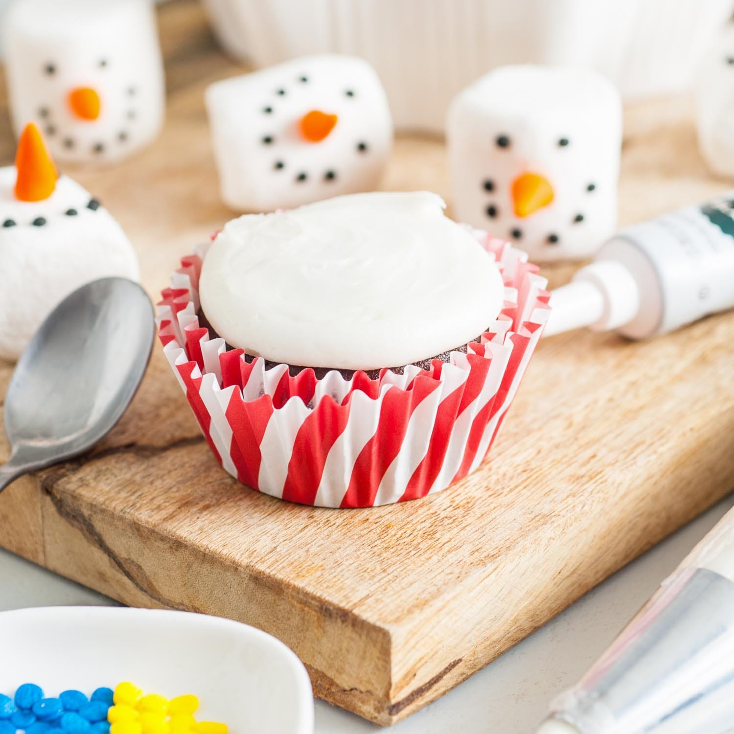 White icing dolloped on top of a chocolate cupcake. In the background are marshmallows decorated to look like the heads of snowmen.