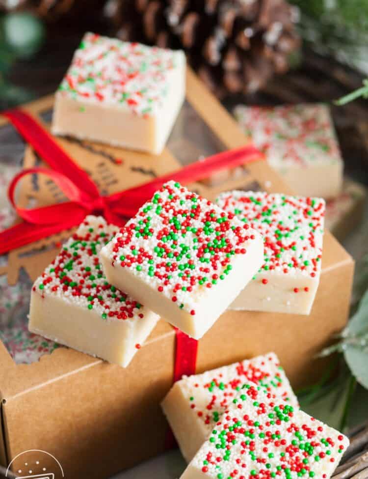 square pieces of holiday sugar cookie fudge on top of a kraft paper gift box that is wrapped with a red velvet ribbon.