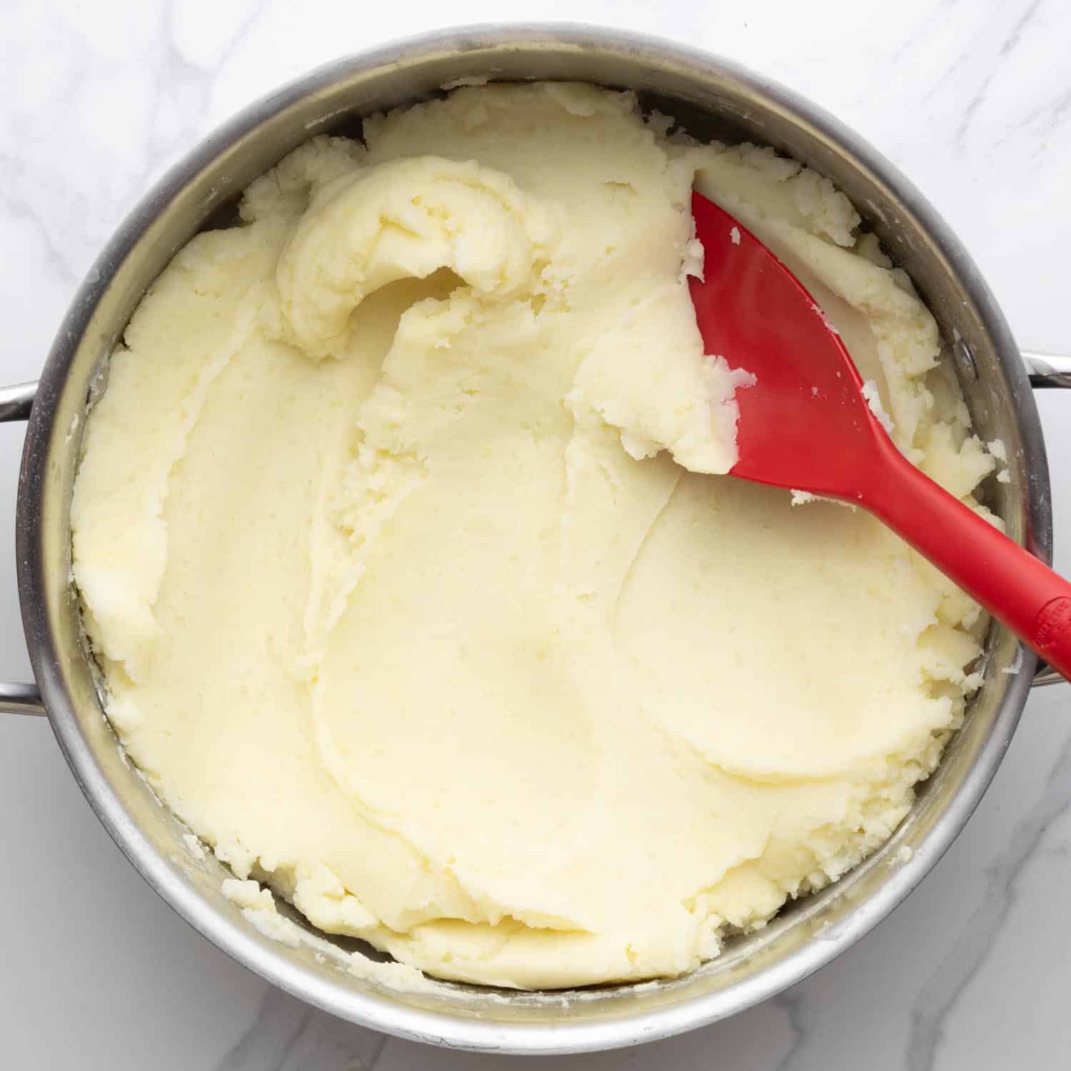 a metal pot of creamy mashed potatoes, stirred with a red spoon.