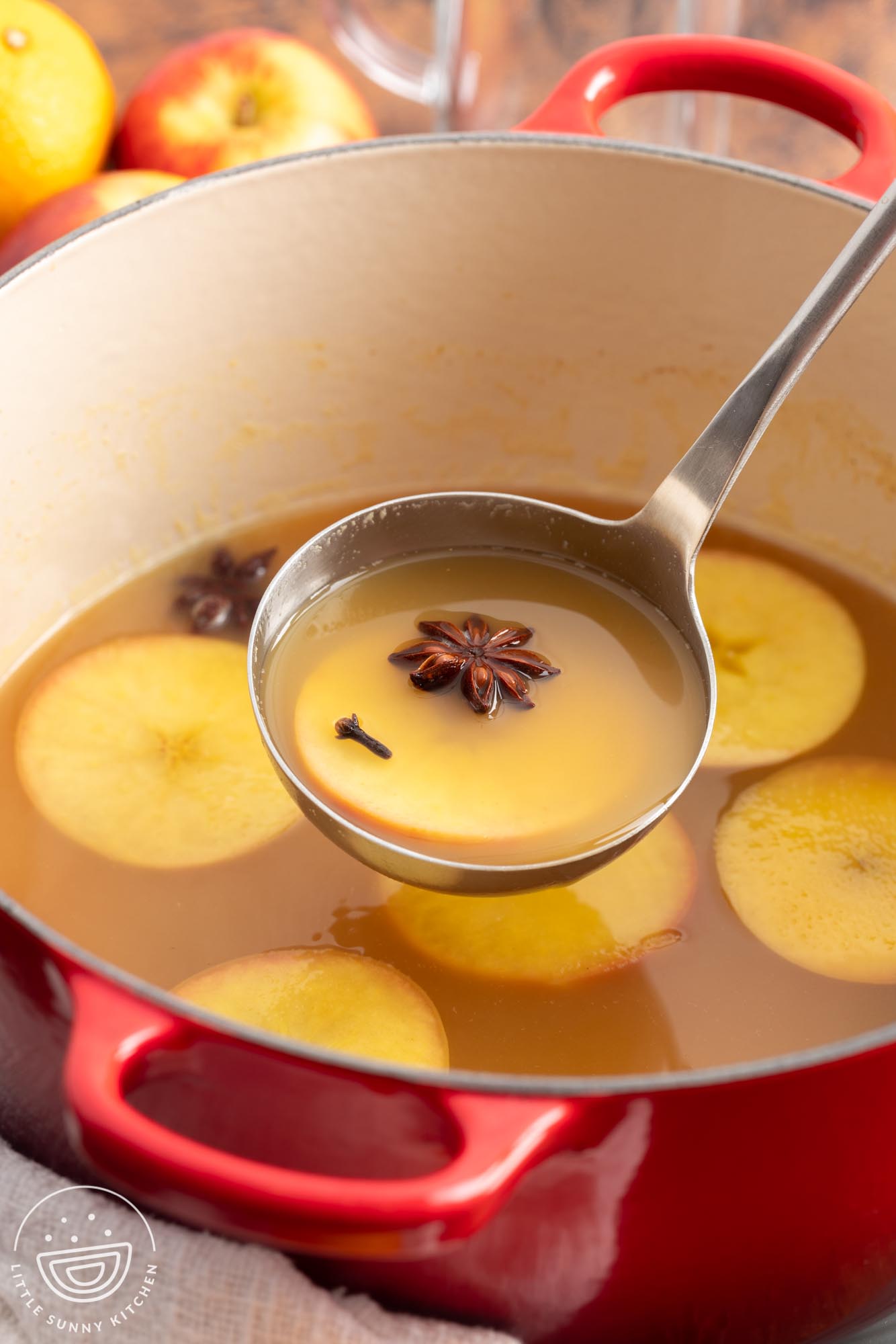 a ladle serving wassail from a red pot, including sliced apples, anise pods, allspice.