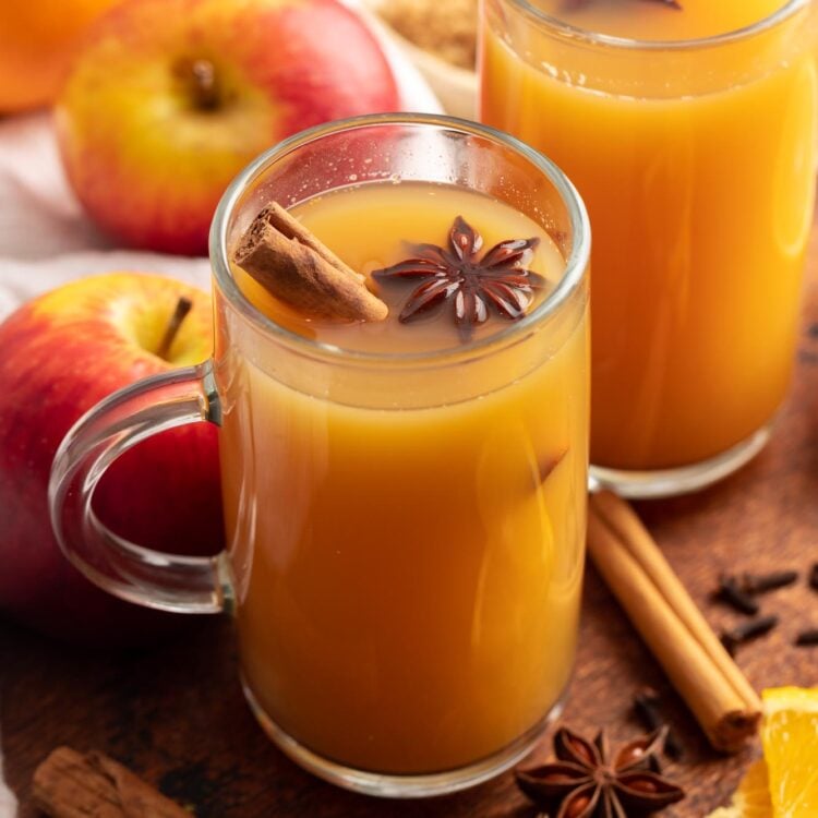 Two mugs of homemade wassail with cinnamon sticks. Around the mugs are fresh apples and oranges and whole spices.