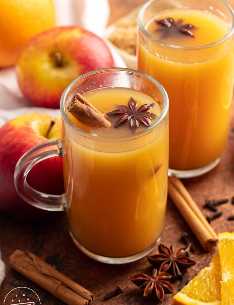 Two mugs of homemade wassail with cinnamon sticks. Around the mugs are fresh apples and oranges and whole spices.