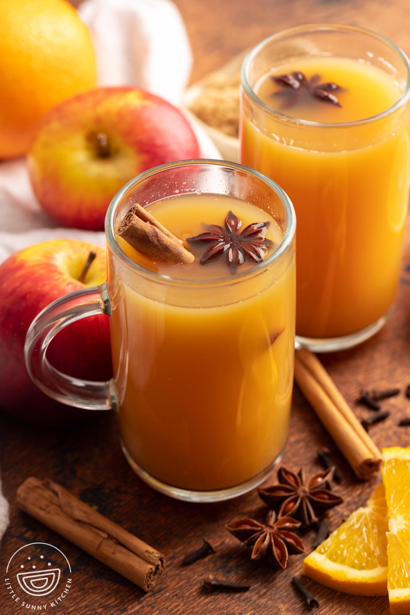 Two mugs of homemade wassail with cinnamon sticks. Around the mugs are fresh apples and oranges and whole spices.