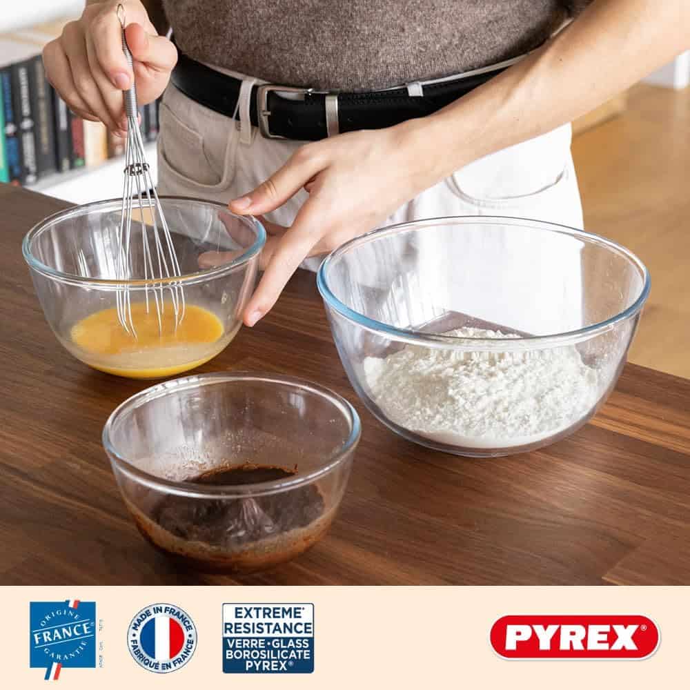 Person whisking ingredients in a Pyrex glass mixing bowl on a wooden countertop. Two additional Pyrex bowls contain dry and liquid ingredients. Pyrex branding and certification icons, including 'Made in France' and 'Extreme Resistance Borosilicate Glass,' are displayed at the bottom of the image