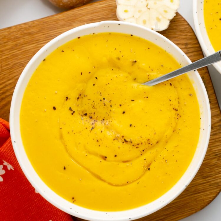 a white bowl of acorn squash soup on a wooden board next to a loaf of bread.