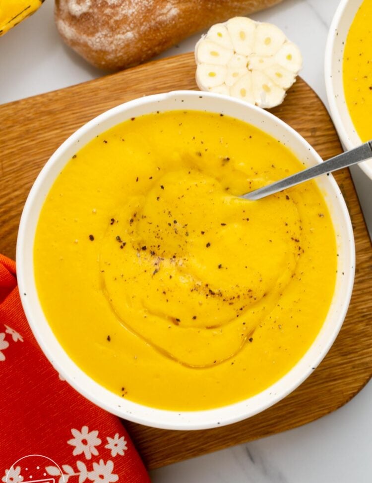 a white bowl of acorn squash soup on a wooden board next to a loaf of bread.