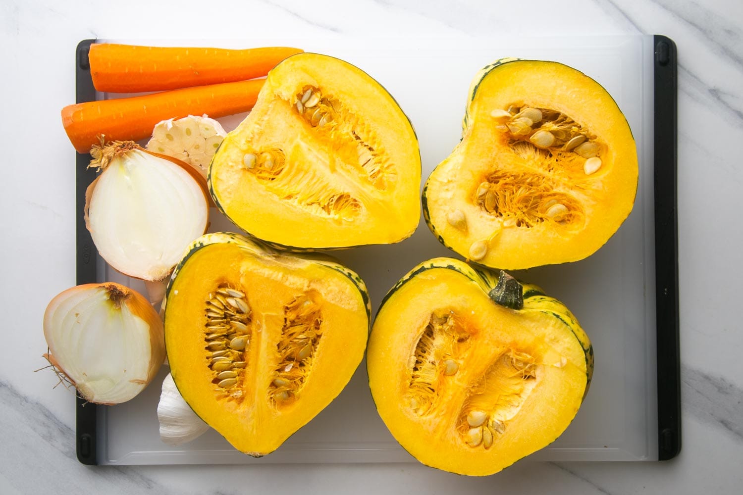 Two acorn squash, cut in half. One onion, cut in half, one head of garlic, cut in half, and two peeled carrots on a cutting board.