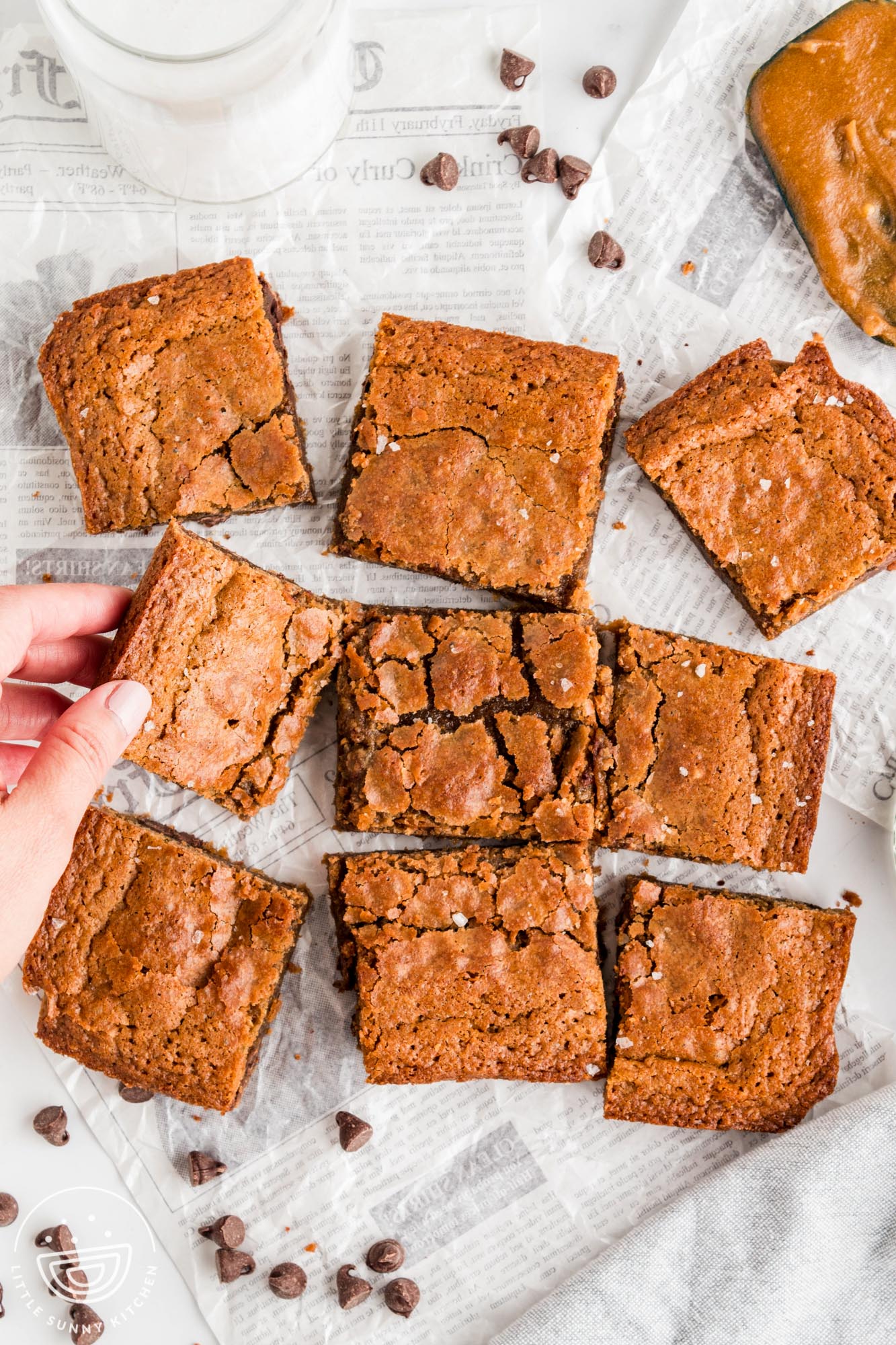 overhead view of a pan of brown butter blondies cut into squares and arranged on a table with a glass of milk. A hand is picking one up.