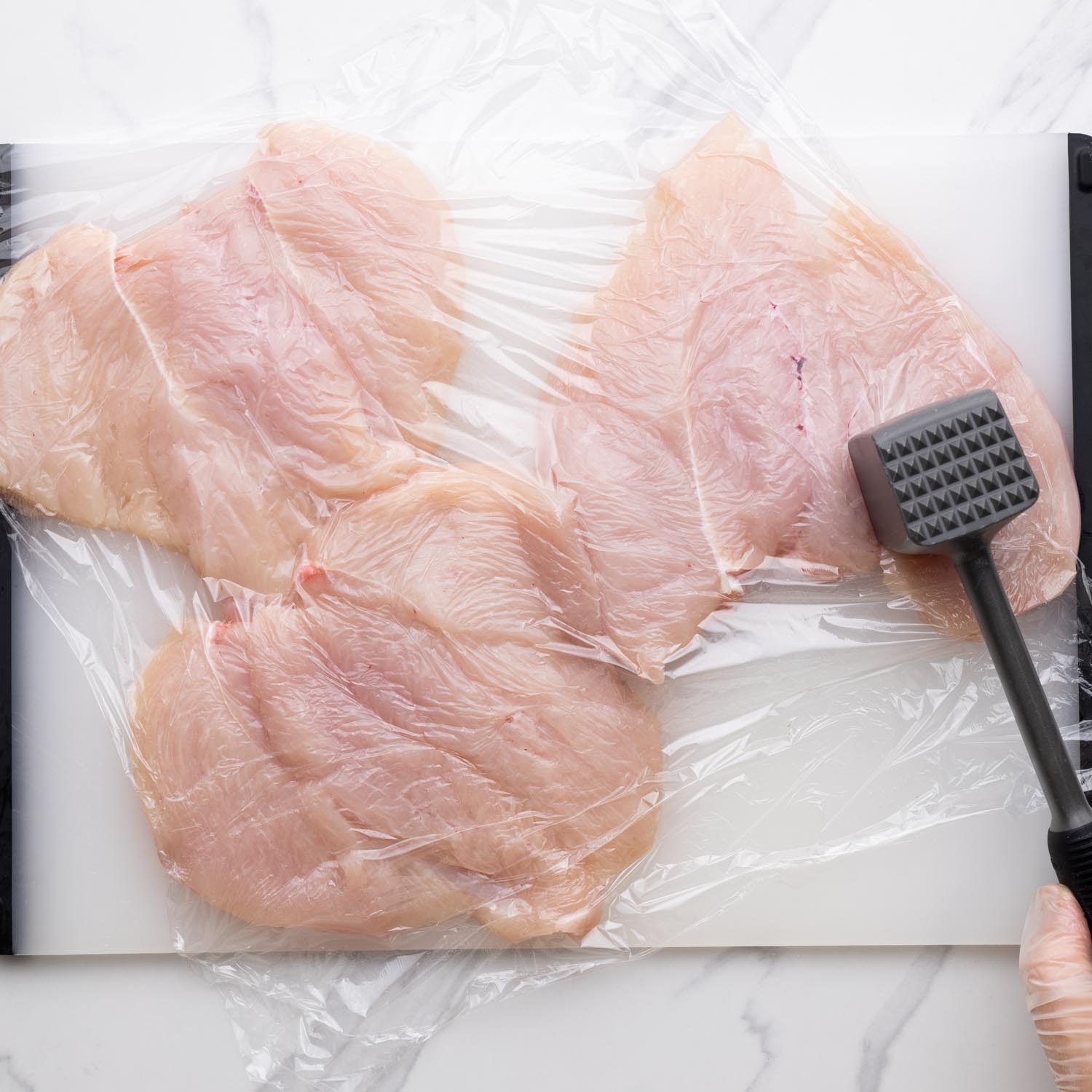 three chicken breasts under plastic wrap, pounded with a meat mallet.