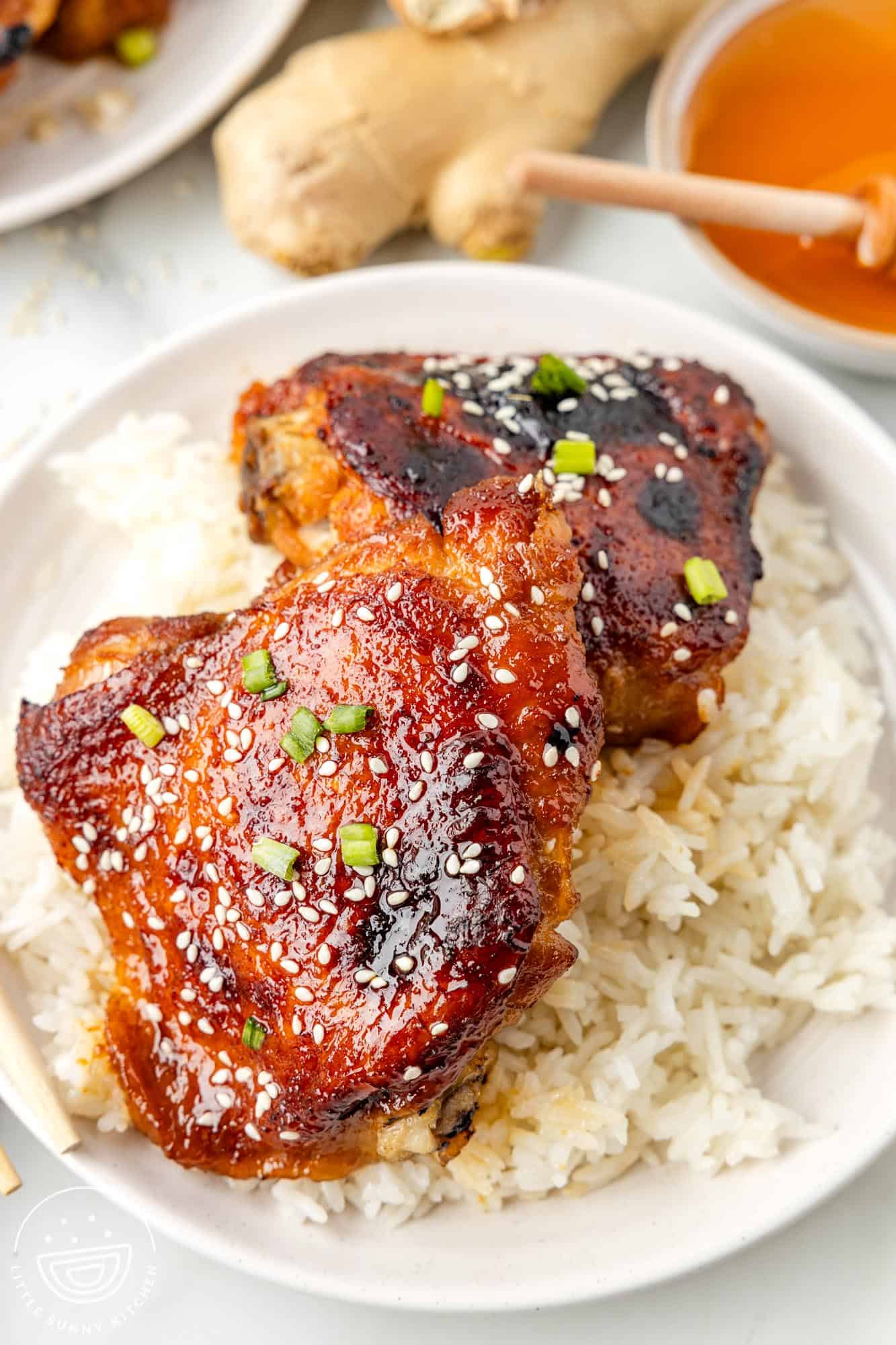 two honey soy chicken thighs on a plate of rice. Behind the plate is a ginger root and a small bowl of honey.