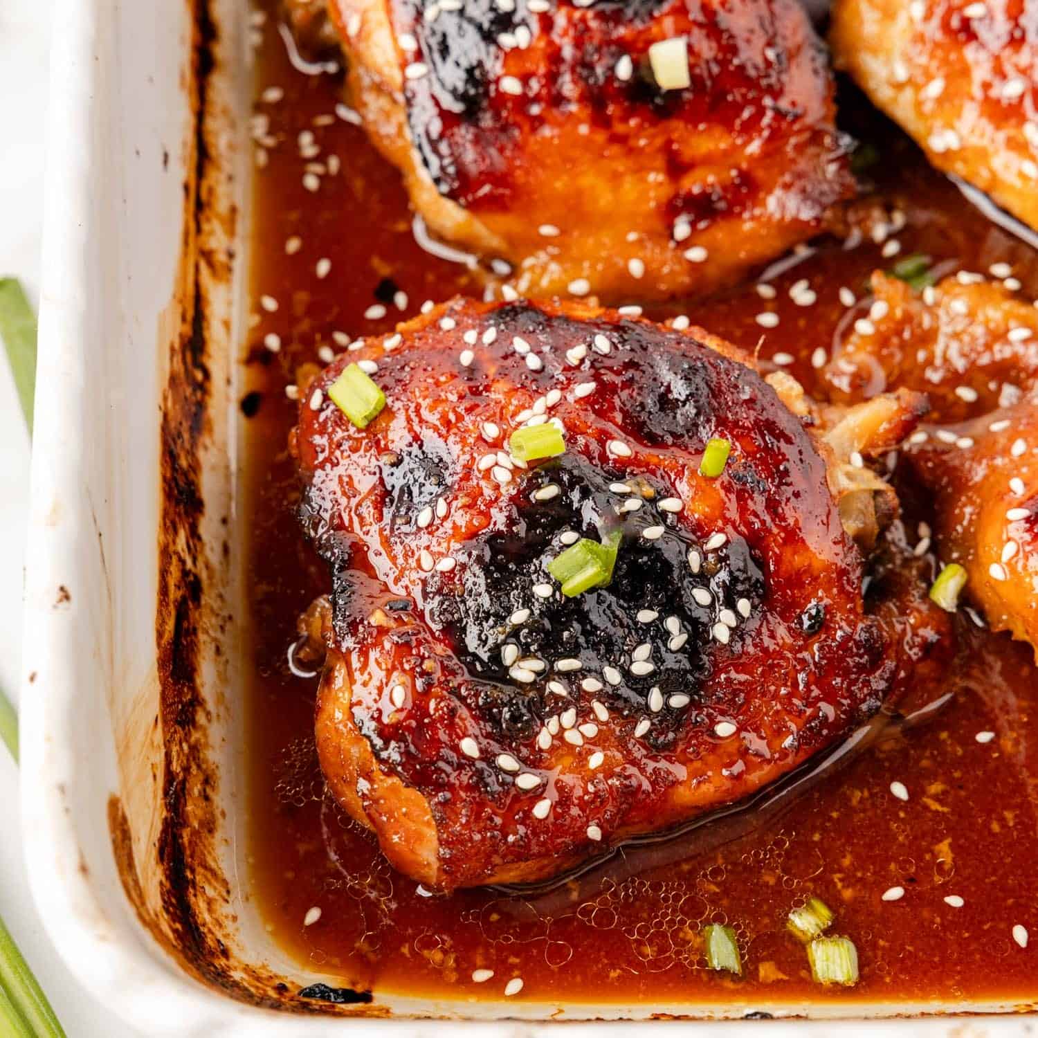 browned and baked honey soy chicken thighs in a white p an, garnished with sesame seeds and green onion.