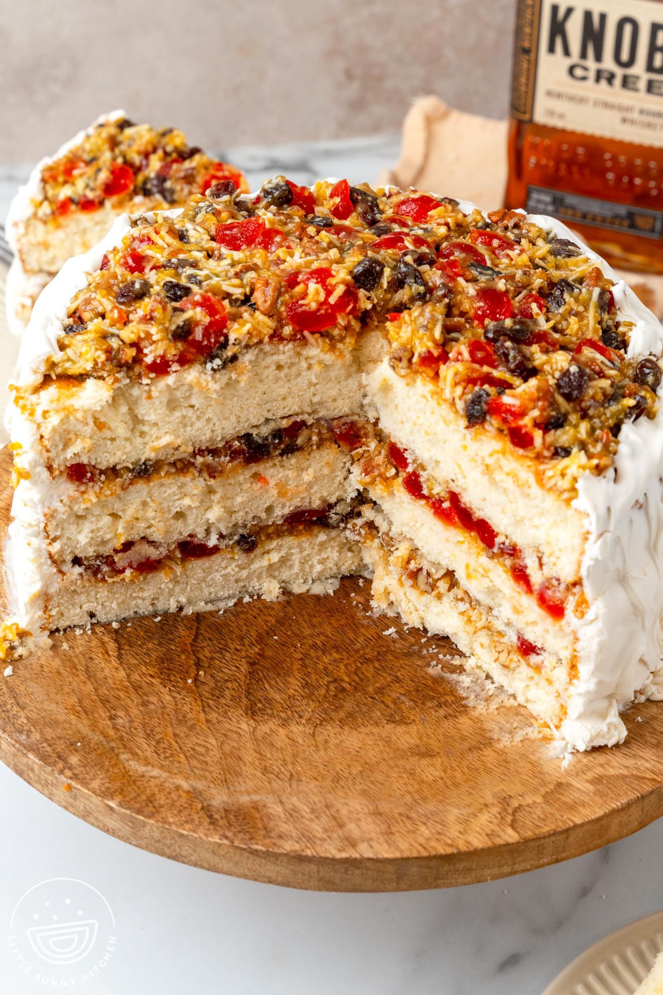 Three layer lane cake with coconut and fruit filling and fluffy frosting. In the background there is a bottle of knob creek boubon.