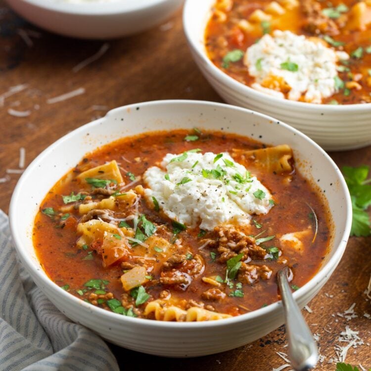 two white bowls of lasagna soup with a dollop of ricotta cheese and other cheeses, garnished with fresh herbs.