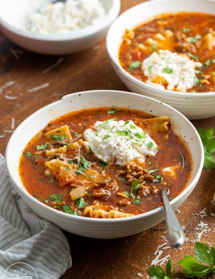 two white bowls of lasagna soup with a dollop of ricotta cheese and other cheeses, garnished with fresh herbs.