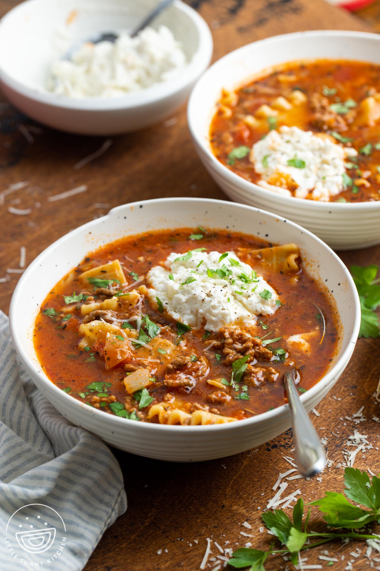 two white bowls of lasagna soup with a dollop of ricotta cheese and other cheeses, garnished with fresh herbs.