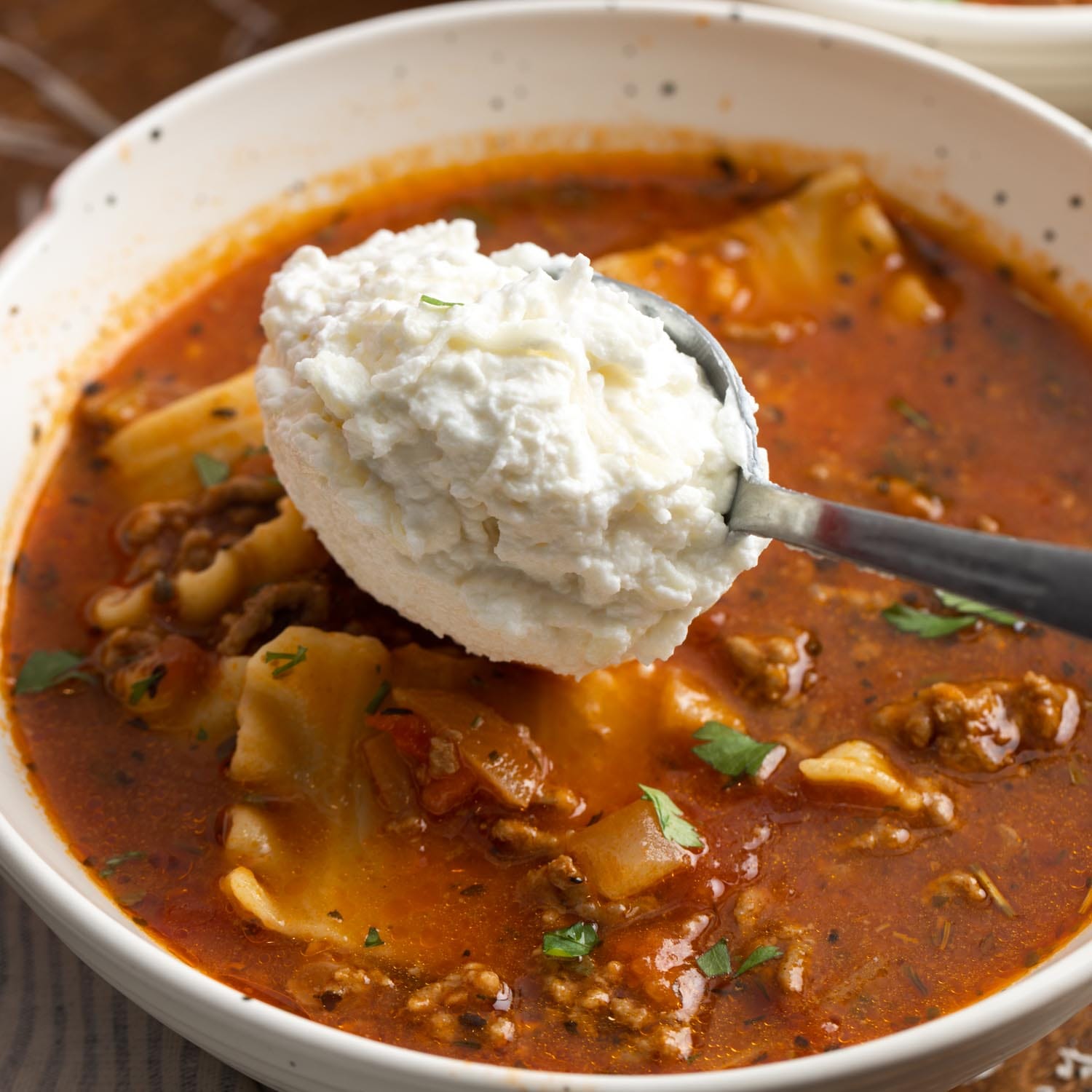 a spoon adding ricotta cheese to a bowl of lasagna soup.