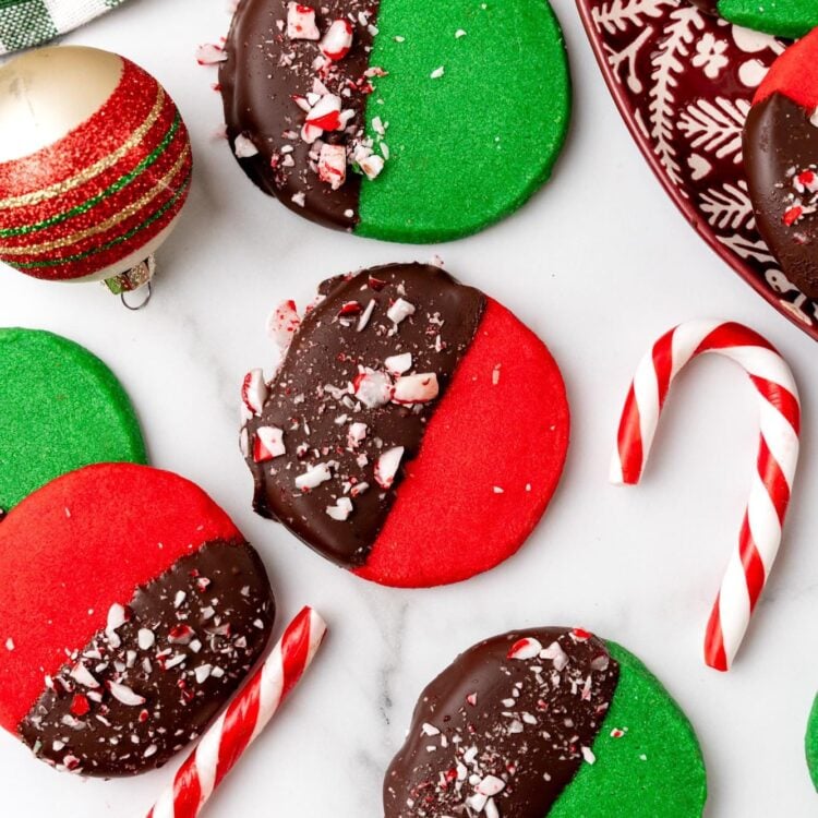 deep red and green round shortbread cookies. Each cookie is half dipped in chocolate and topped with crushed peppermint candy. The cookies are arranged on a marble counter with candy canes and christmas ornaments.