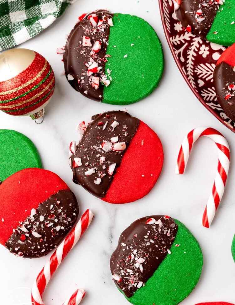 deep red and green round shortbread cookies. Each cookie is half dipped in chocolate and topped with crushed peppermint candy. The cookies are arranged on a marble counter with candy canes and christmas ornaments.