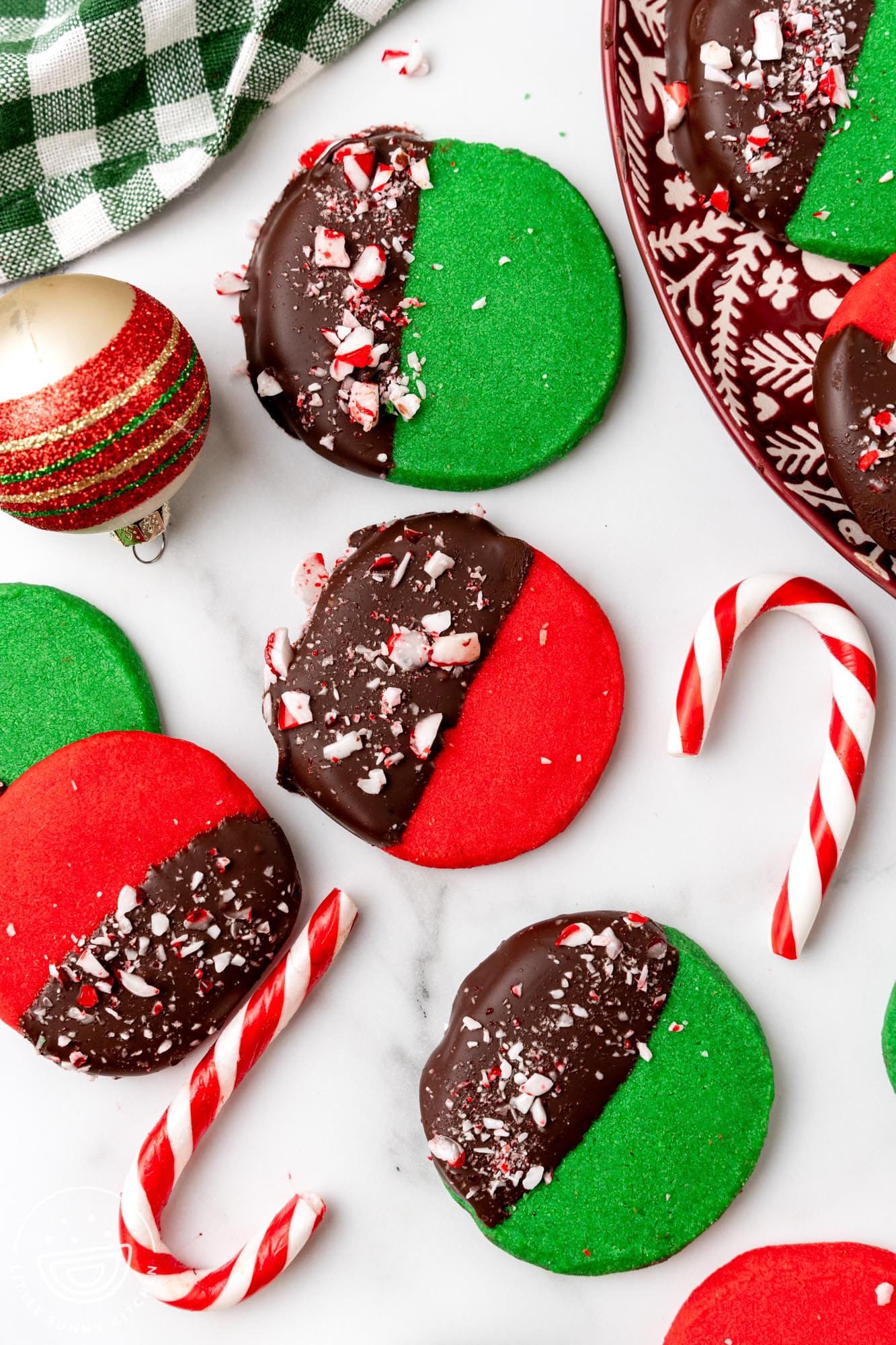 deep red and green round shortbread cookies. Each cookie is half dipped in chocolate and topped with crushed peppermint candy. The cookies are arranged on a marble counter with candy canes and christmas ornaments.