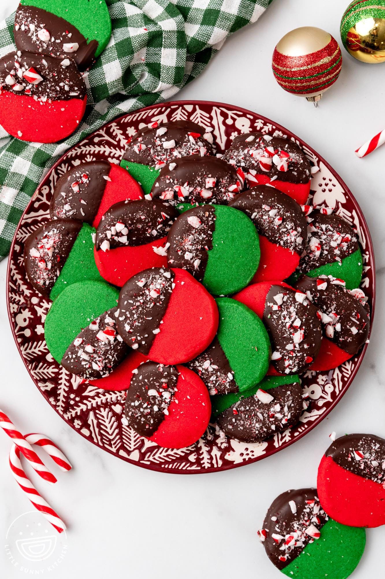 a plate of christmas colored peppermint shortbread cookies with chocolate coating. Around the plate are candy canes and holiday ornaments.