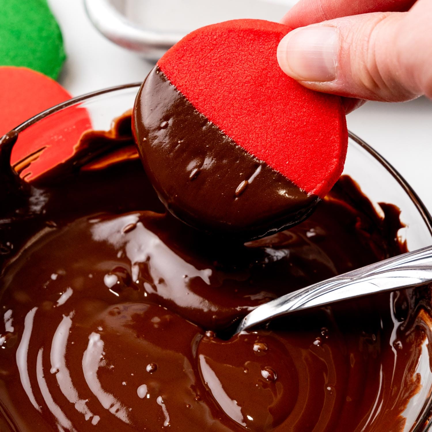 a hand dipping a round red cookie into melted chocolate.