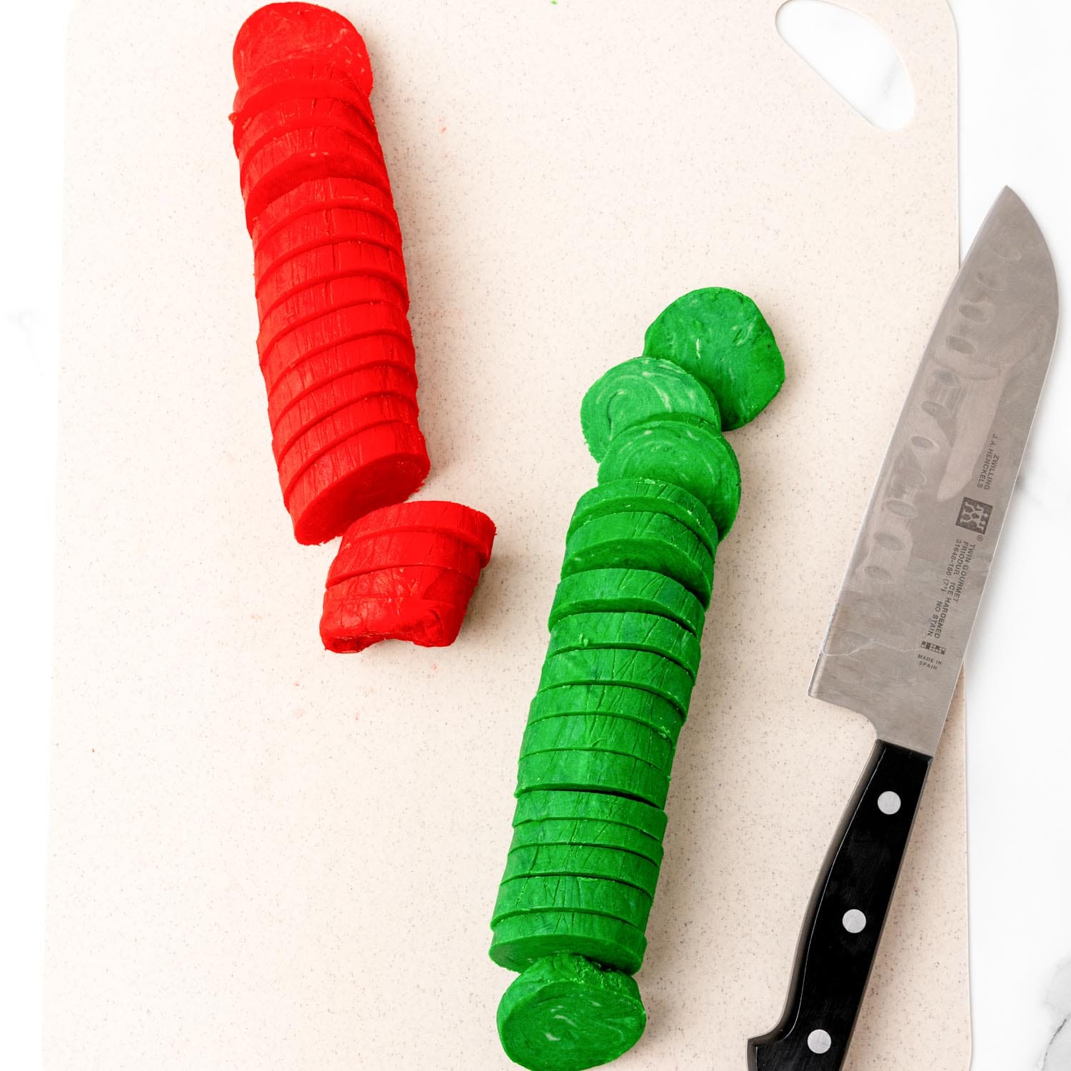 logs of red and green cookie dough, sliced into rounds with a chefs knife on a cutting board.