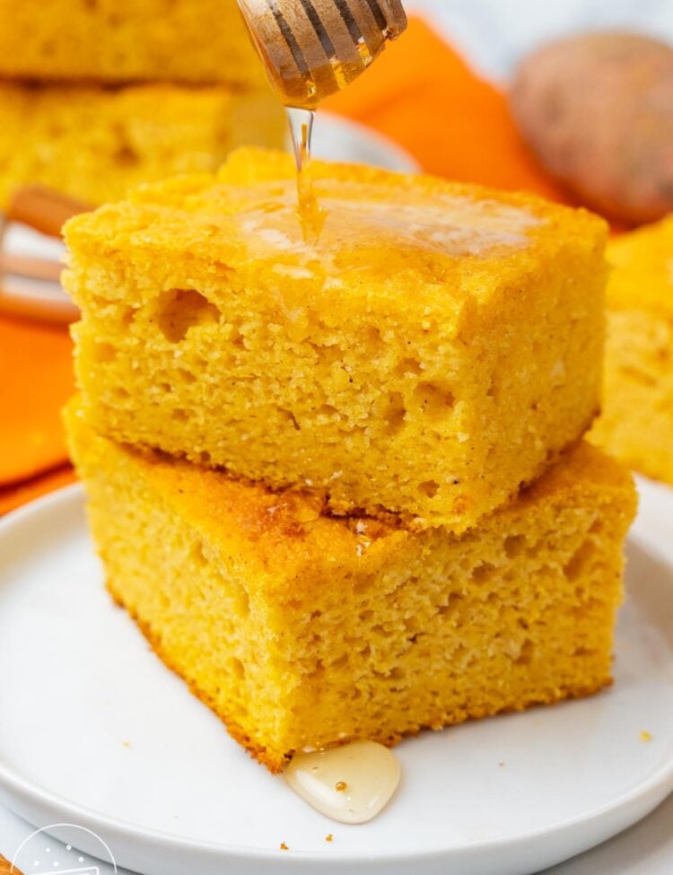 two square pieces of sweet potato cornbread on a plate. Honey is drizzled over.