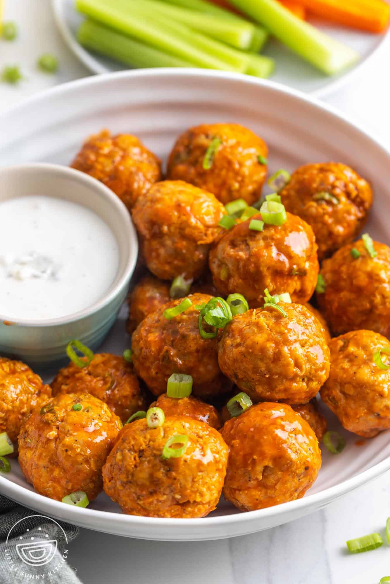 a plate of chicken meatballs with buffalo sauce, garnished with green onion. With the meatballs is a cup of blue cheese dressing for dipping.