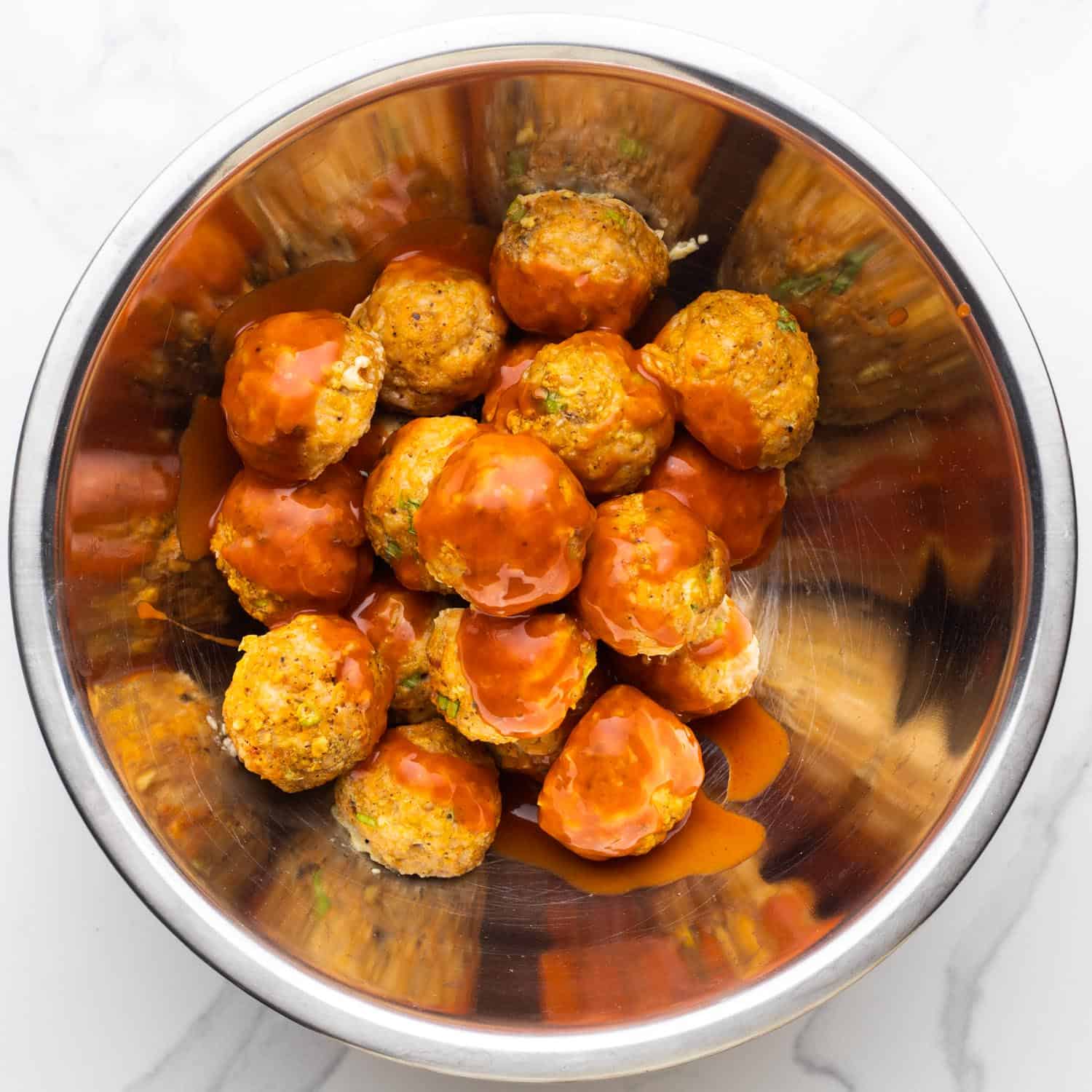 wing sauce added to buffalo chicken meatballs in a metal mixing bowl.