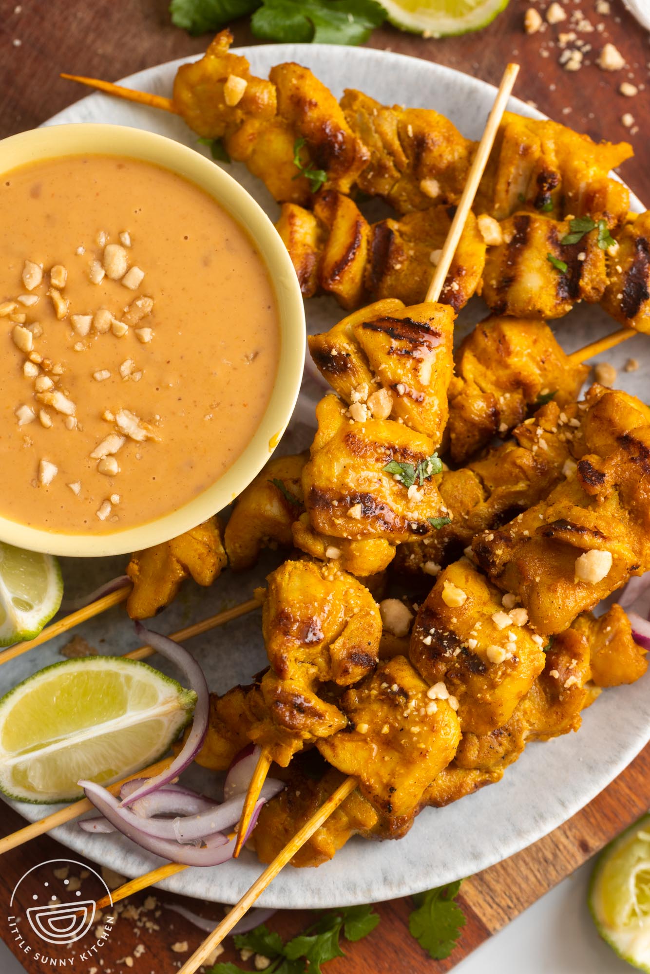 a large plate of grilled chicken satay skewers with a bowl of peanut sauce. On the side are lime wedges and red onion.