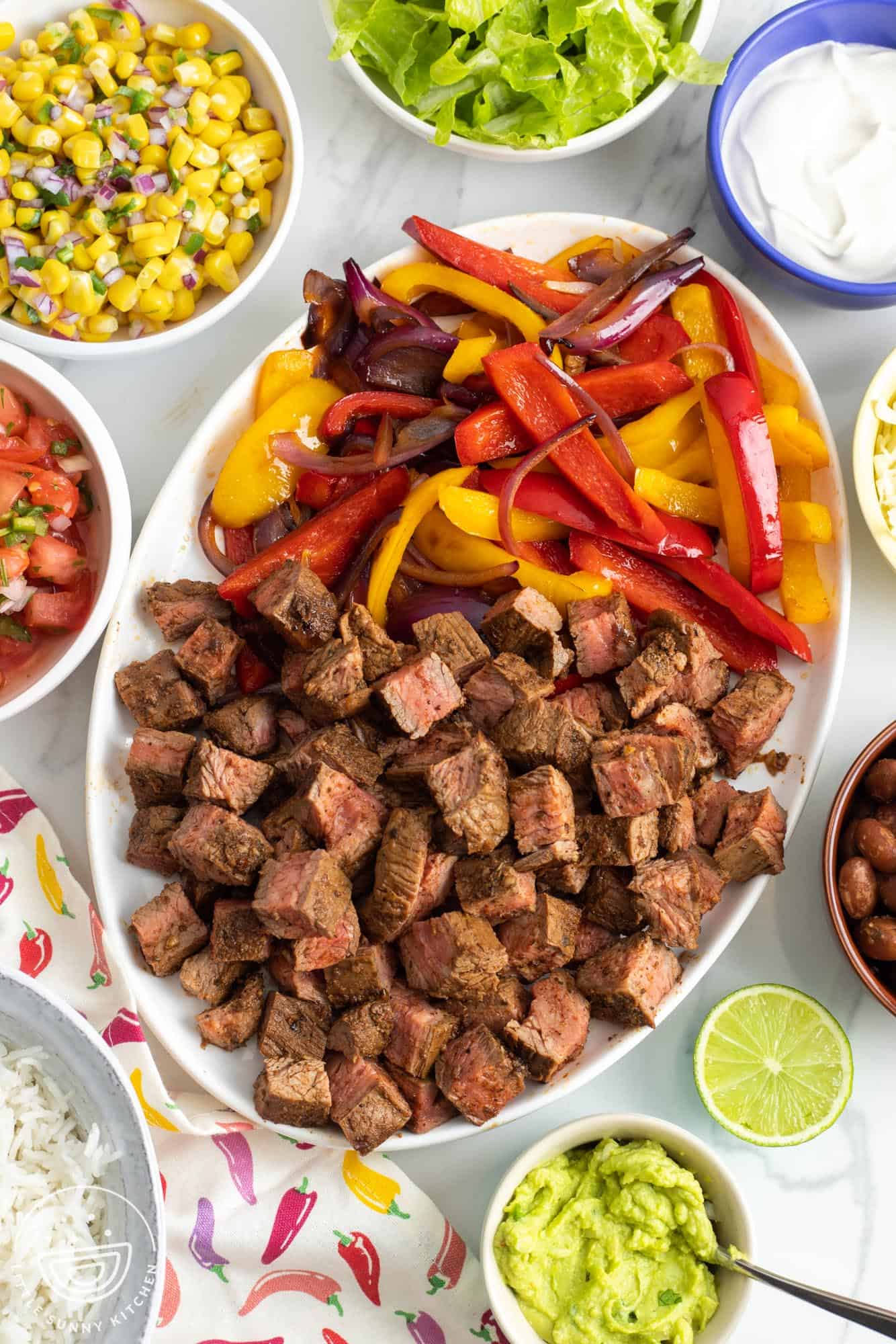 a platter of chipotle steak and peppers and onions. Around the platter are small bowls of toppings, including corn salsa, lettuce, sour cream, and guacamole.