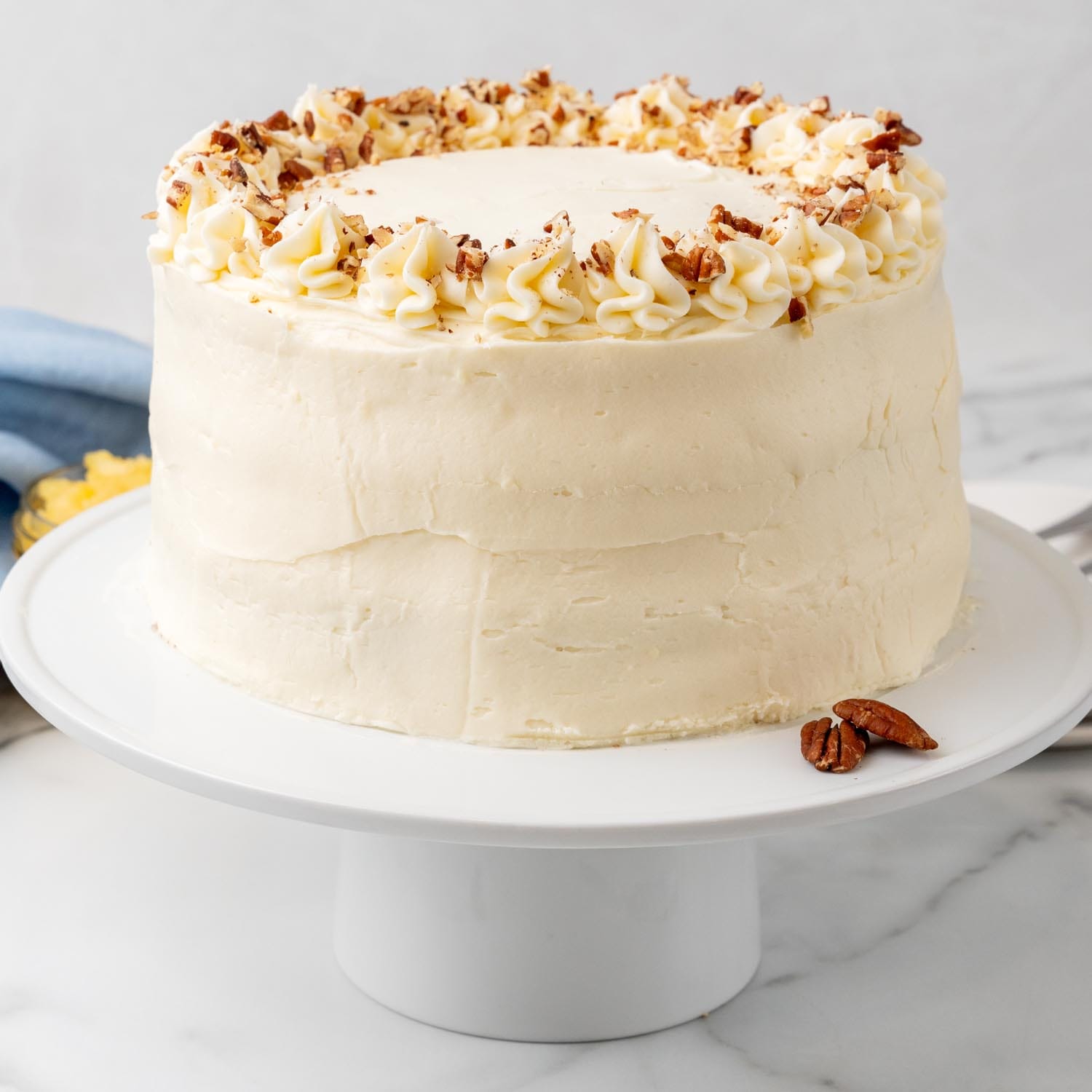 a white cake pedestal holding a tall round hummingbird cake, iced, topped with icing details and crushed pecans.